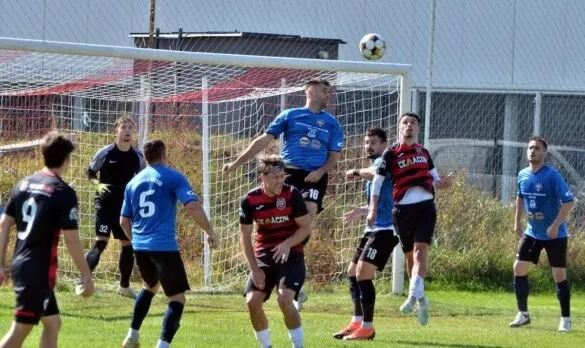 Câțiva jucători de fotbal de pe un teren de iarbă sunt încadrați în fața unei porți de fotbal sub un cer senin.