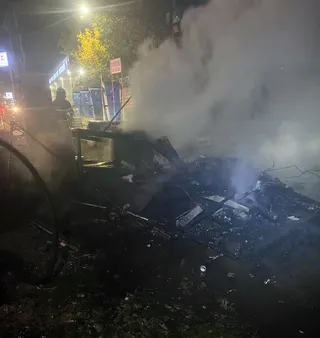 Un vehicul de tip rulotă este acoperit de fum dens și resturi carbonizate pe un teren întunecat noaptea.