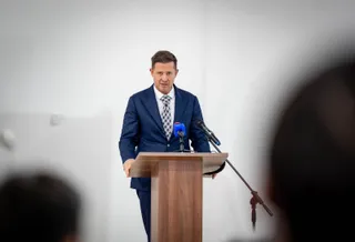Un bărbat într-un costum albastru și cravată stă în spatele unui podium de lemn cu două microfoane în fața unui fundal alb.