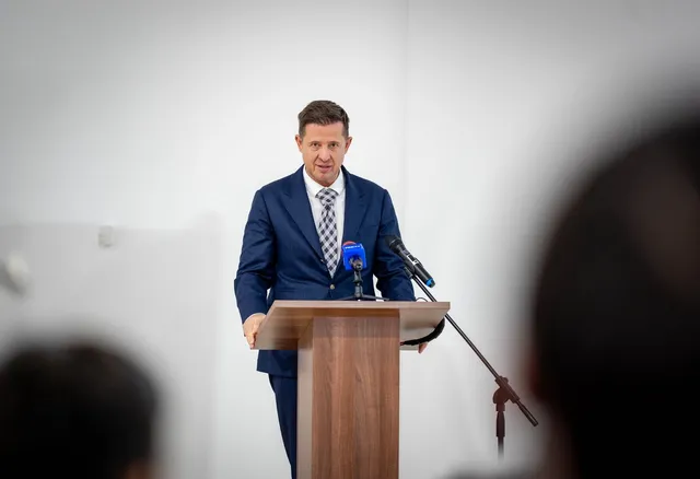 Un bărbat într-un costum albastru și cravată stă în spatele unui podium de lemn cu două microfoane în fața unui fundal alb.
