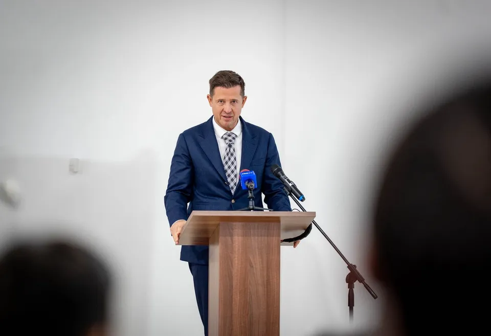 Un bărbat într-un costum albastru și cravată stă în spatele unui podium de lemn cu două microfoane în fața unui fundal alb.