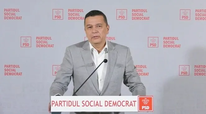 Un bărbat cu părul scurt și sacou gri stă în fața unui fundal alb cu logourile roșii ale Partidului Social Democrat.