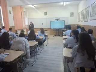 O femeie stă în fața unui grup de elevi așezați la bănci într-o sală de clasă cu un ecran digital pe perete.