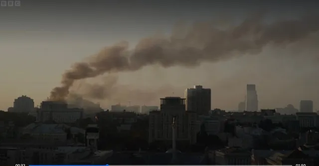 Un nor de fum gros se ridică deasupra unei siluete urbane compuse din diverse clădiri sub un cer de seară.