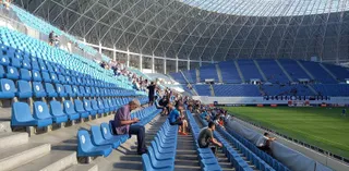 Scaune de culoare albastră sunt dispuse în rânduri pe tribunele unui stadion, unde câțiva spectatori stau așezați.