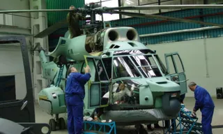 Un grup de tehnicieni în uniforme albastre lucrează la un elicopter de culoare verde deschis aflat într-un hangar.