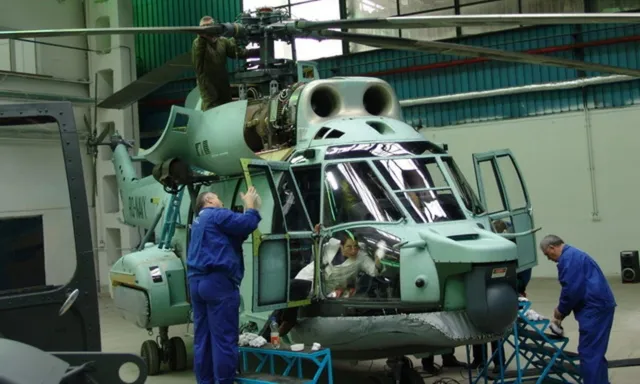 Un grup de tehnicieni în uniforme albastre lucrează la un elicopter de culoare verde deschis aflat într-un hangar.