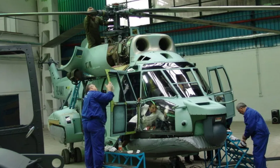 Un grup de tehnicieni în uniforme albastre lucrează la un elicopter de culoare verde deschis aflat într-un hangar.