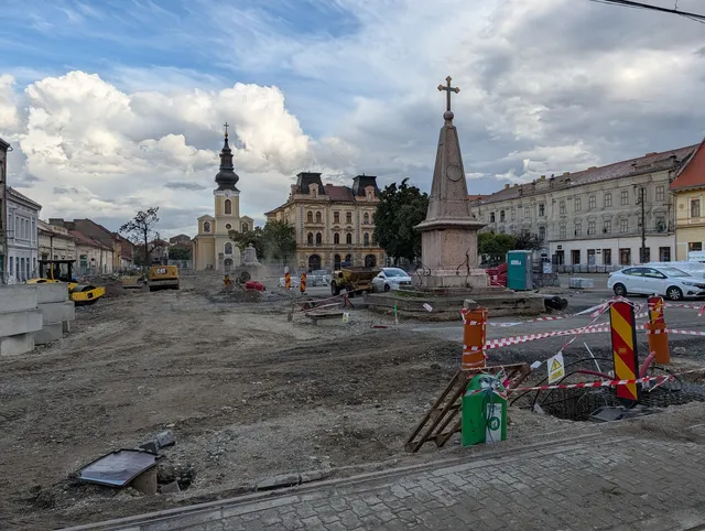 Un șantier cu pământ descoperit, utilaje de construcții, un monument cu o cruce și clădiri vechi sub un cer cu nori.