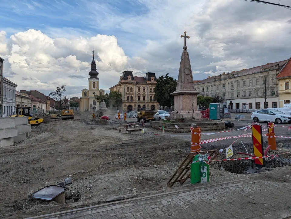 Un șantier cu pământ descoperit, utilaje de construcții, un monument cu o cruce și clădiri vechi sub un cer cu nori.