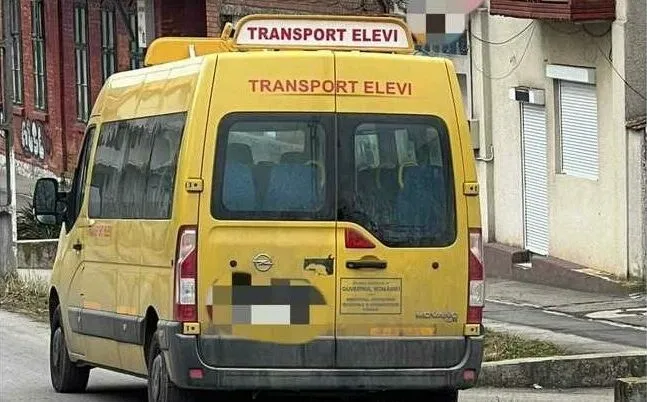 Un microbuz galben cu inscripții de transport elevi este parcat pe un drum lângă o clădire cu ferestre și jaluzele.