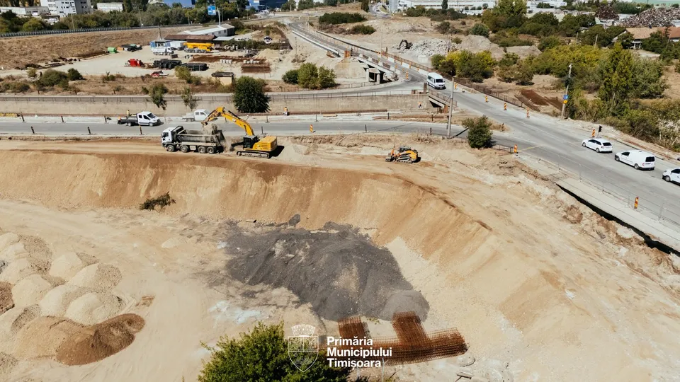 O excavatoare galbenă lucrează lângă un camion alb pe un șantier de construcții cu pământ și nisip.