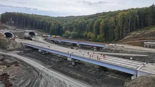 Două structuri de pod din beton cu suprafață gri trec peste un șantier de construcții înconjurat de dealuri împădurite sub un cer înnorat.