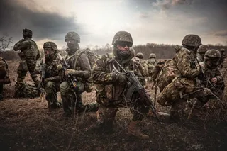 Soldați în uniforme de camuflaj și echipament militar stau ghemuiți pe un teren împădurit sub un cer înnorat.
