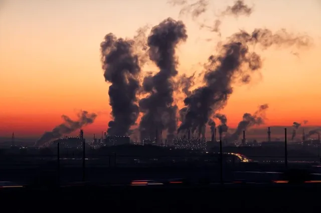 Siluete de coloane de fum negru se ridică deasupra unei zone industriale sub un cer la apus.