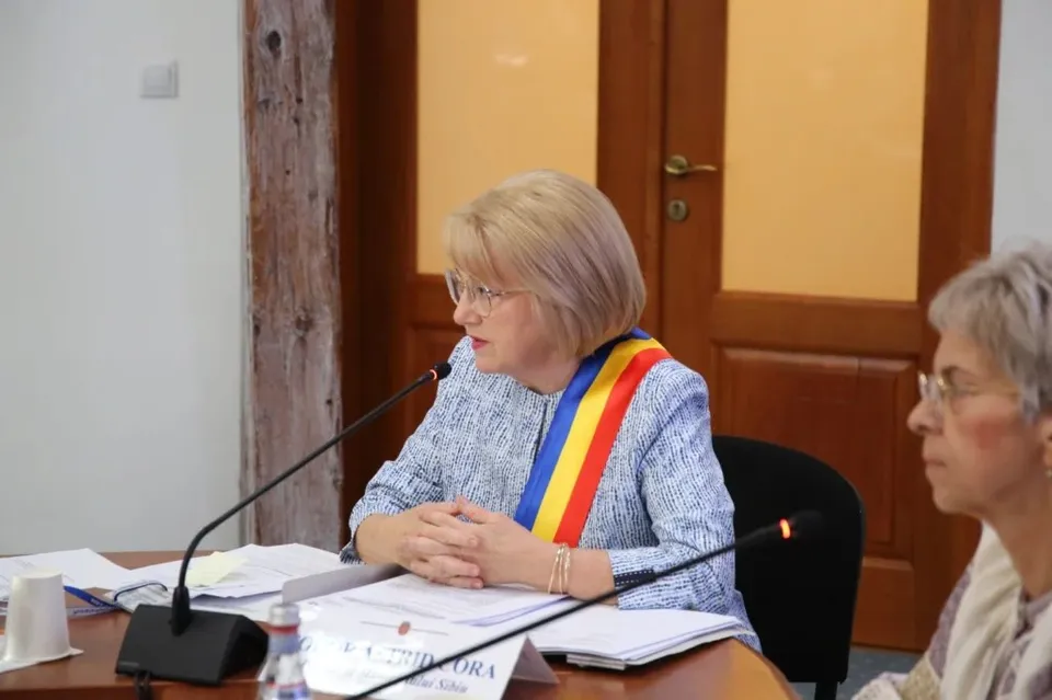 O femeie cu părul scurt și ochelari poartă o panglică tricoloră peste o bluză albastră și stă la o masă cu microfoane.