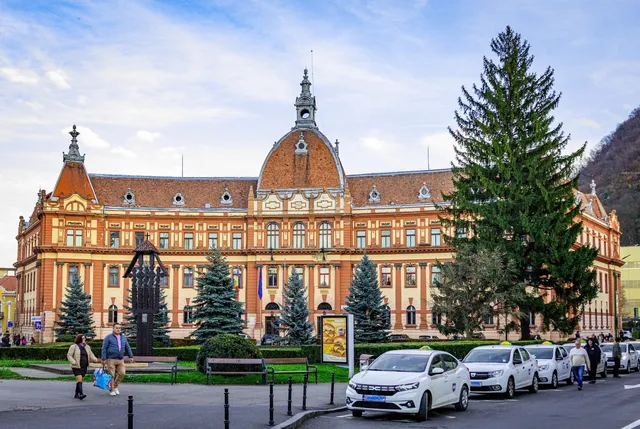 O clădire mare, de culoare portocalie cu detalii albe, este situată în spatele unor arbori coniferi și a unor mașini parcate pe un drum asfaltat.