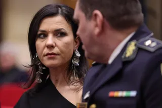 O femeie cu părul negru și cercei argintii mari privește spre un bărbat purtând o uniformă în culori închise cu epoleți și insignee.