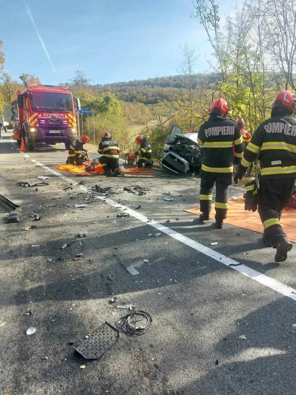 Pompieri în echipament de protecție portocalie și negru stau pe un drum asfaltat lângă un vehicul de serviciu roșu și resturi împrăștiate.