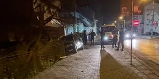Un autoturism negru este poziționat pe marginea trotuarului lângă un gard, în timp ce mai mulți oameni stau pe trotuar lângă un vehicul alb.