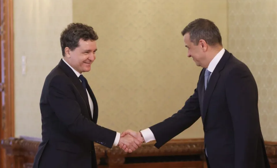 Doi bărbați în costume închise de culoare stau față în față și se strâng de mână într-o încăpere cu pereți deschiși de culoare deschisă.