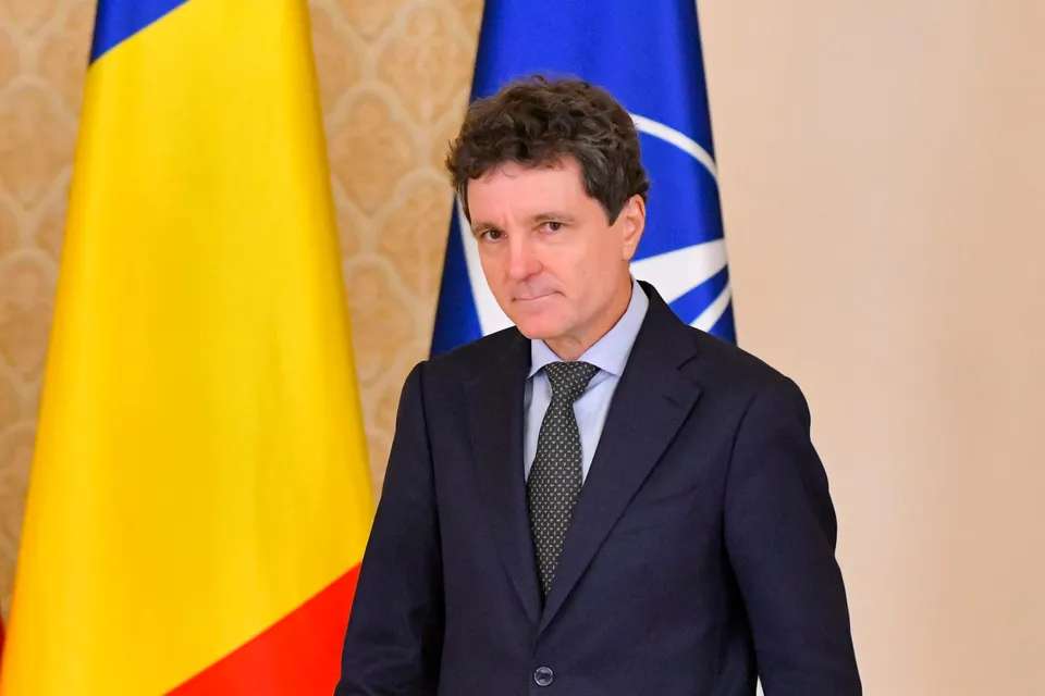 Un bărbat cu părul închis la culoare, purtând costum închis la culoare și cravată, stă în fața unui steag galben, albastru și roșu și a unui steag albastru.
