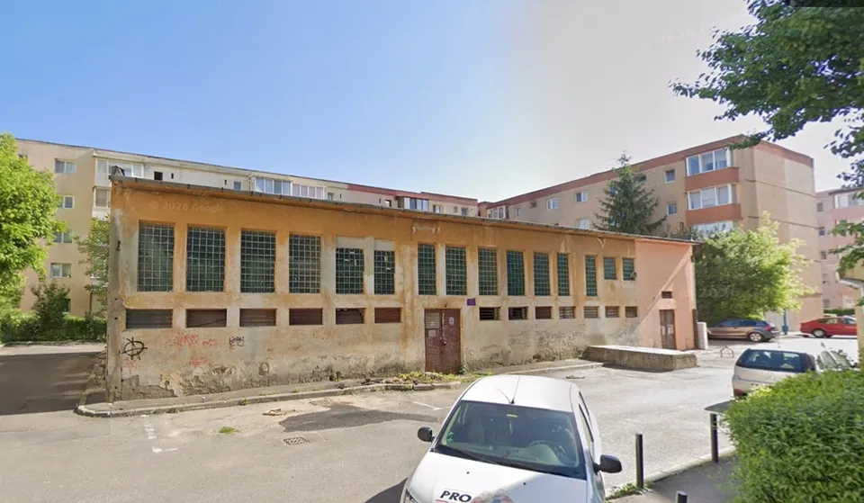 O clădire lungă, de un singur etaj, cu ferestre înalte și ziduri uzate, este situată lângă blocuri de locuințe sub un cer senin.