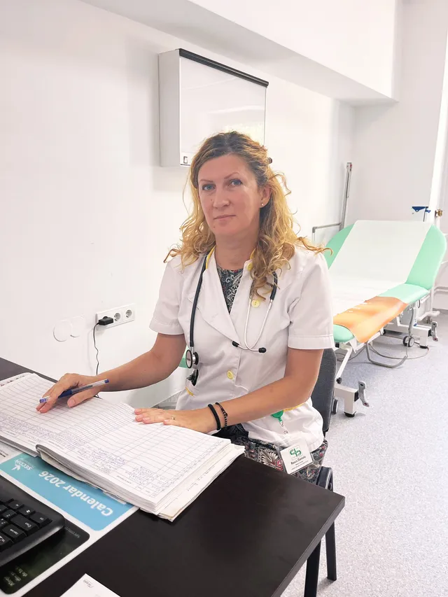 O femeie cu părul blond și halat alb stă la un birou cu un registru deschis și un stetoscop în jurul gâtului într-o cameră medicală.