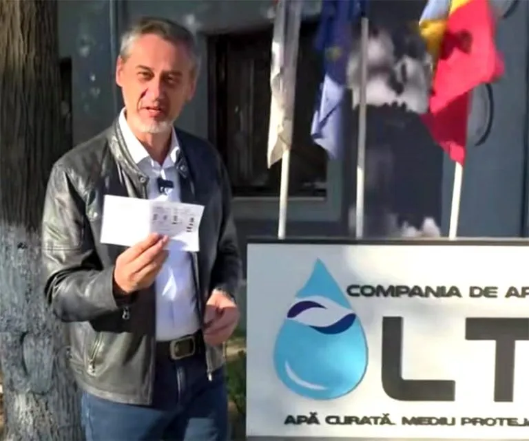 Un bărbat cu geacă de piele și cămașă albă ține o foaie de hârtie în fața unui panou informativ cu un logo albastru.