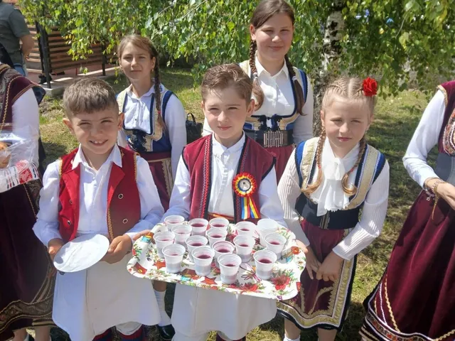 Patru fete în costume populare tradiționale stau în picioare lângă o tavă cu pahare mici cu lichid roșu.