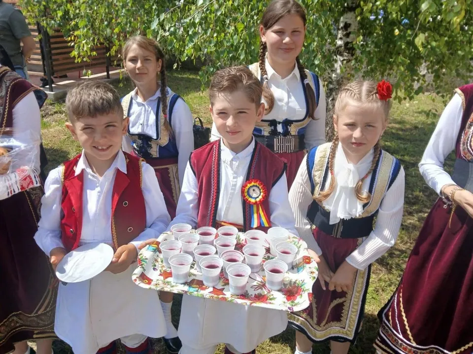 Patru fete în costume populare tradiționale stau în picioare lângă o tavă cu pahare mici cu lichid roșu.