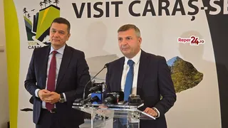 Doi bărbați în costume și cravate stau în fața unui panou publicitar cu text și imagini de peisaje.