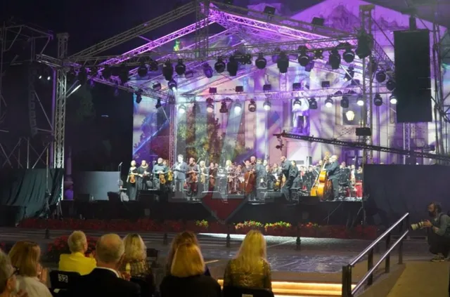 Un ansamblu de muzicieni cu instrumente de coardă execută un spectacol pe o scenă iluminată cu lumini mov în fața unui public așezat.