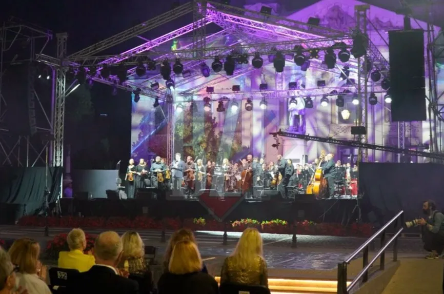Un ansamblu de muzicieni cu instrumente de coardă execută un spectacol pe o scenă iluminată cu lumini mov în fața unui public așezat.