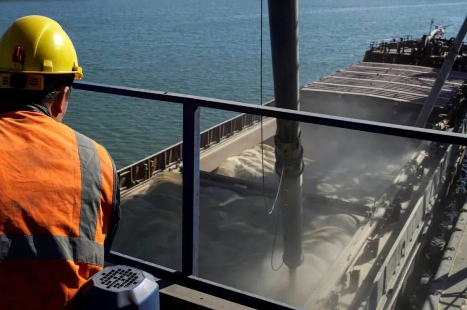 Un bărbat cu cască galbenă și vestă reflectorizantă privește un vas de marfă care descarcă un material granular într-un compartiment.