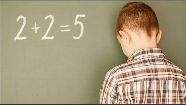 Un copil cu o cămașă în croit este așezat în fața unei table verzi pe care este scris cu cretă ecuația matematică 2+2=5.