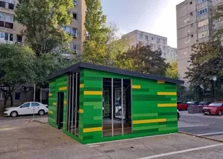O structură mică, rectangulară, cu pereți verzi și dungi galbene, este amplasată pe un teren asfaltat lângă clădiri de locuințe și copaci.