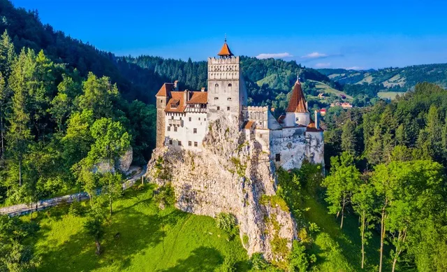 O cetate medievală cu turnuri înalte și acoperișuri în pante este situată pe o stâncă înconjurată de păduri verzi sub un cer albastru.