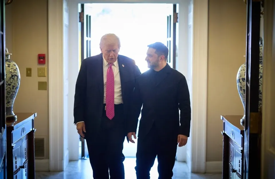 Donald Trump, într-un costum închis cu cravată roz, și Volodimir Zelenski, într-o ținută neagră, mergând alăturat într-un coridor luminos.