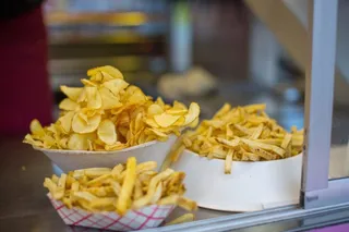 Două boluri de carton conțin cartofi prăjiți și chipsuri de cartofi, așezate pe o suprafață metalică într-un stand de prezentare.