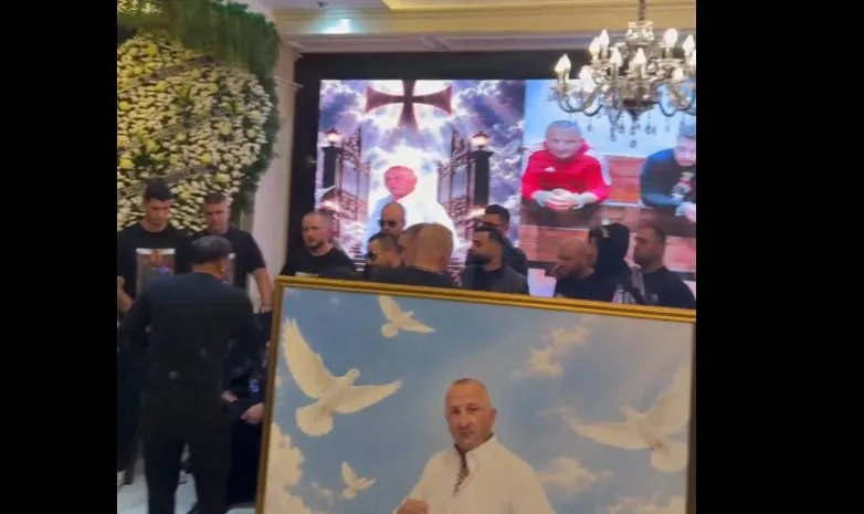 Un grup de bărbați stă în fața unui ecran mare care afișează imagini cu o cruce și un om, lângă un cadru cu porți albe.