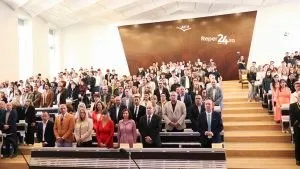 Un grup mare de persoane stă în șaipte în interiorul unei săli de conferințe cu pereți de culoare maro și scări din lemn.
