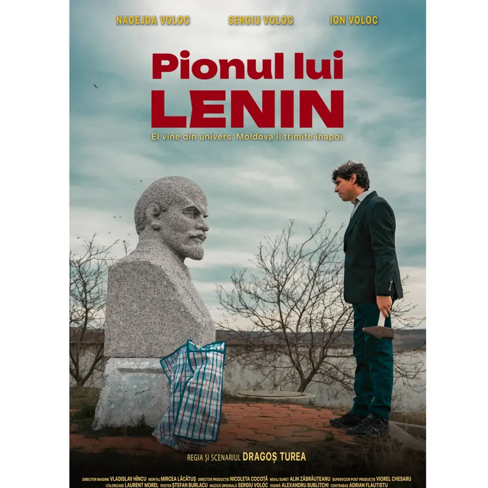 Un bărbat într-un costum închis stă lângă o bustă de piatră gri sub un cer înnorat.
