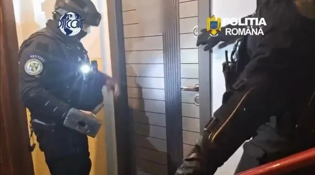 Două persoane în uniforme întunecate cu echipament tactic stau lângă o ușă de interior.