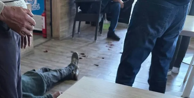 Un bărbat stă întins pe podeaua de lemn, lângă mese și scaune, în timp ce alți indivizi stau în picioare în jurul său.