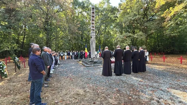 Un grup de persoane stă în picioare pe un teren împădurit lângă un monument înalt și subțire, în jurul căruia se află cruci roșii.