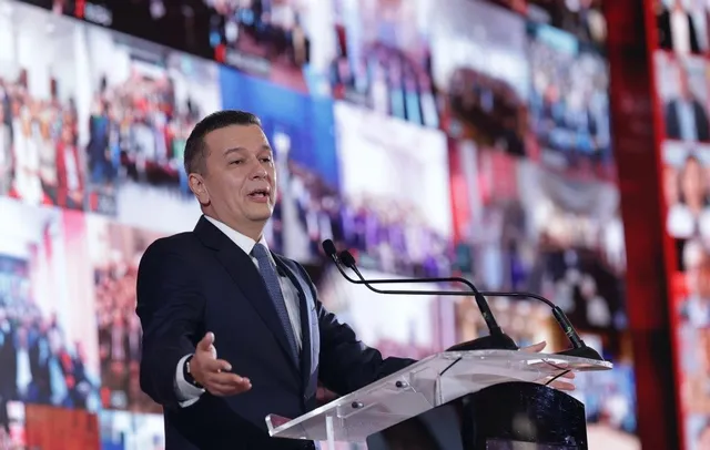 Un bărbat într-un costum închis la culoare și cravată stă în spatele unui podium transparent cu microfoane, în fața unui ecran mare cu imagini.