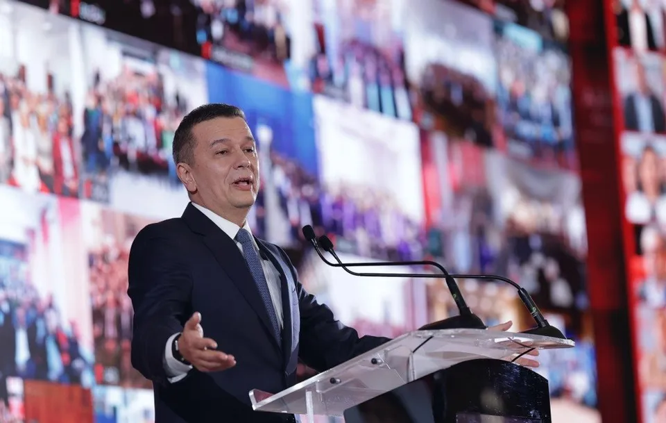Un bărbat într-un costum închis la culoare și cravată stă în spatele unui podium transparent cu microfoane, în fața unui ecran mare cu imagini.