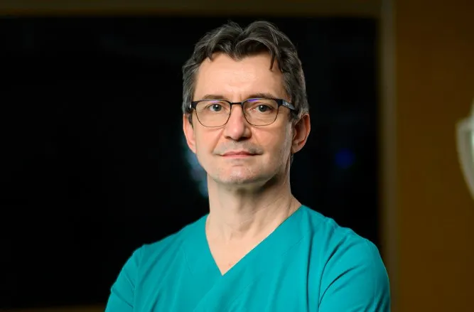 Un bărbat cu ochelari și halat medical de culoare turcoaz privește spre cameră pe un fundal întunecat.