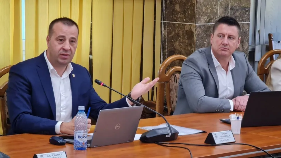 Un bărbat într-un costum albastru stă la o masă de lemn lângă un laptop și un microfon, gestuind cu mâna stângă.